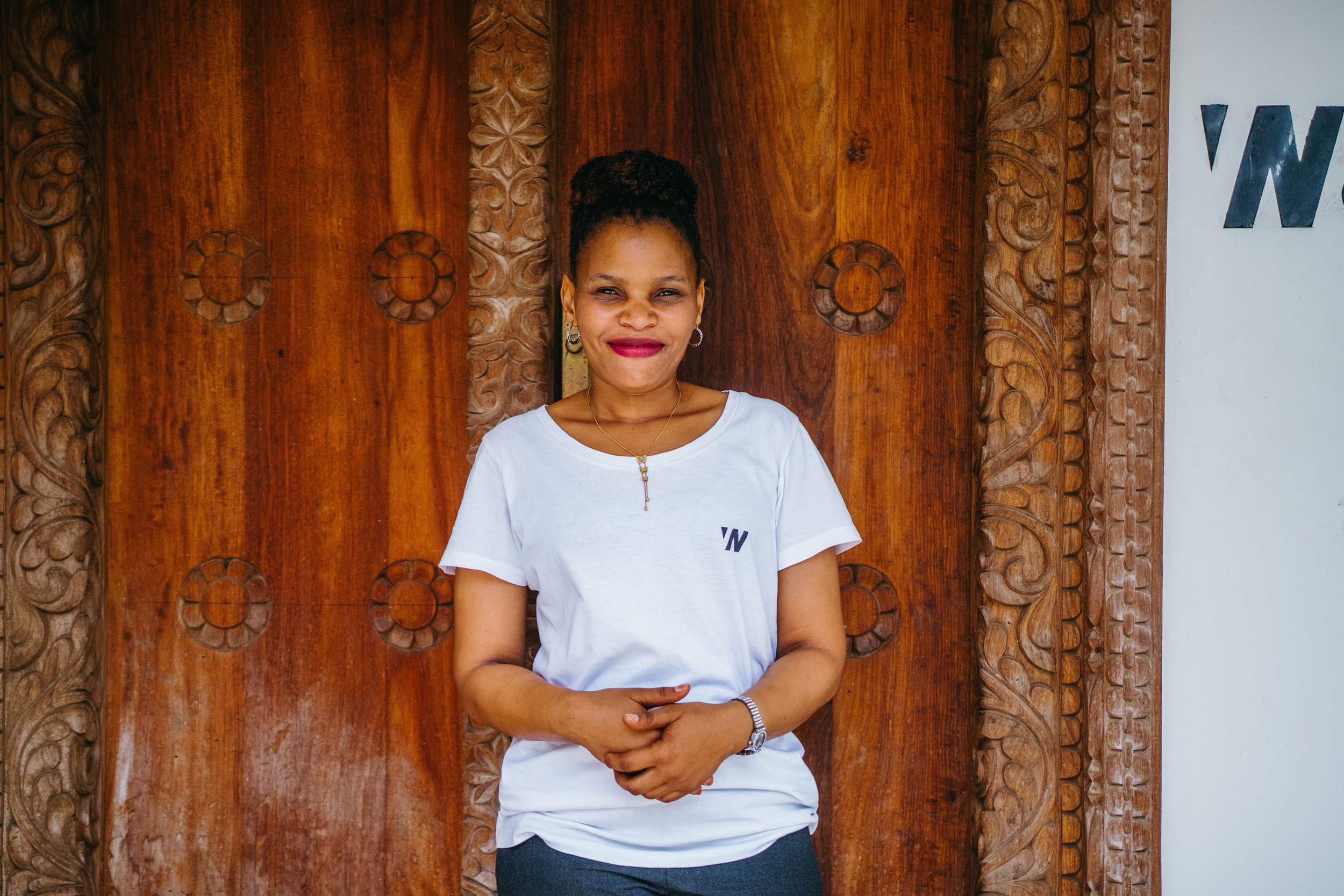 A woman standing in front of a wooden carved door, smiling with her arms crossed, wearing a white t-shirt and jewelry.