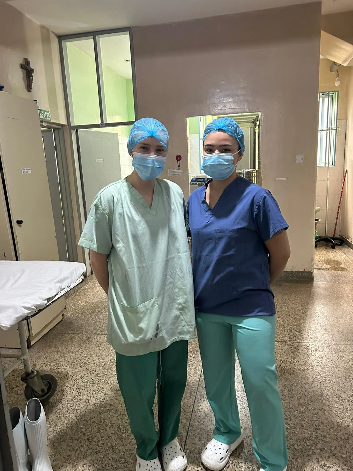 Two healthcare workers in scrubs, masks, and hair caps standing in a hospital hallway.