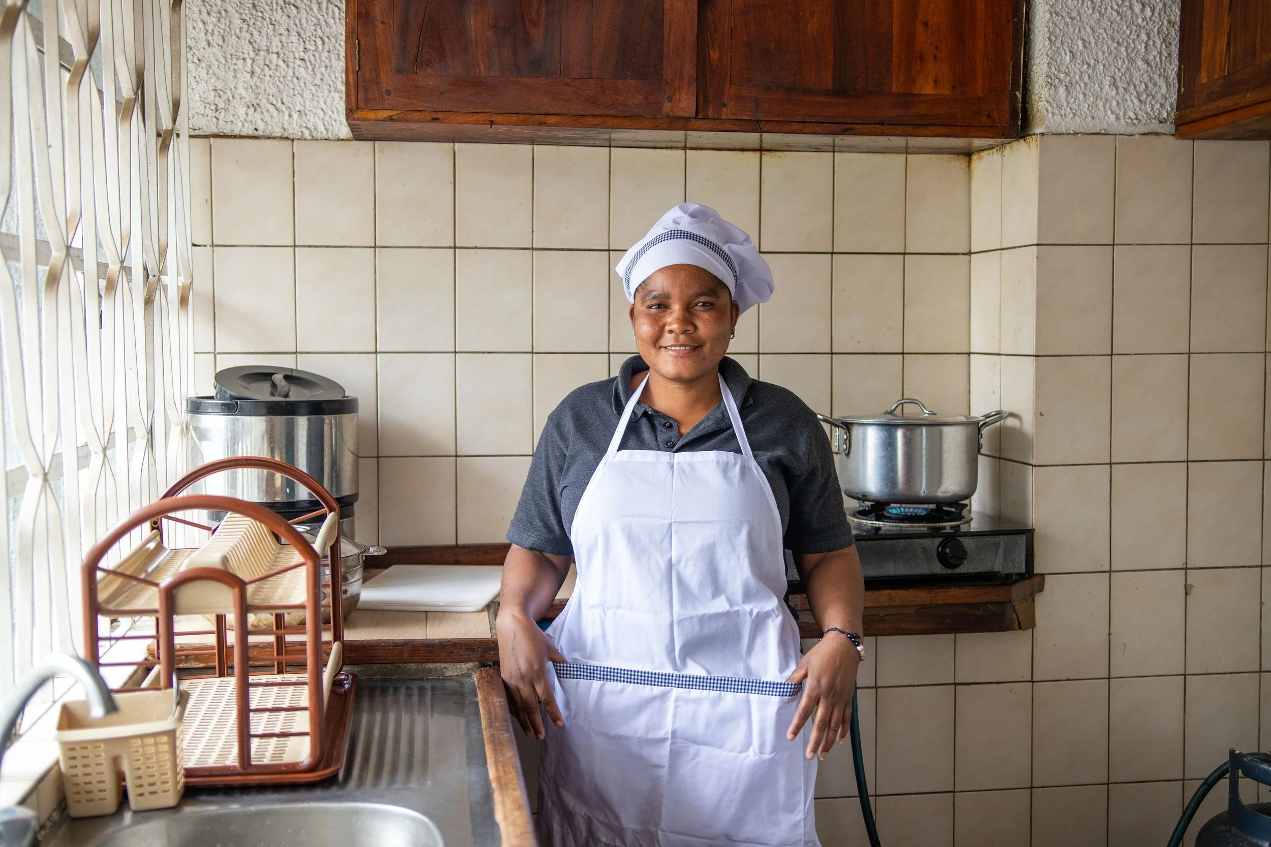 A woman wearing a white chef's hat and apron stands in a kitchen with tiled walls, smiling at the camera. There are pots on the stove behind her and kitchen utensils on the counter in front.
