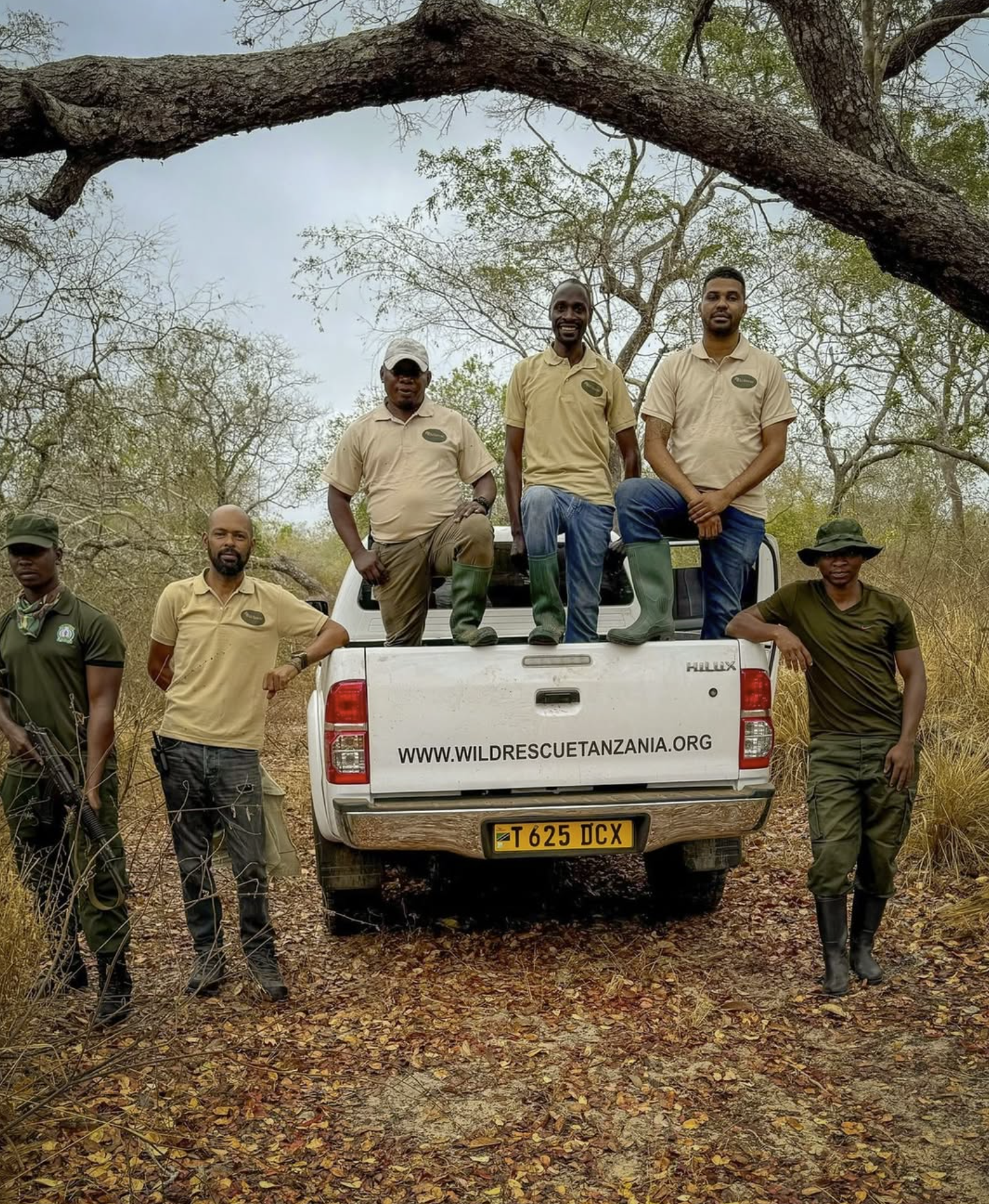 Group of people standing and sitting on a white pickup truck in a dry forested area. The truck has a website URL and a license plate from Tanzania. The individuals are dressed in safari or field work attire, some with boots and hats, indicating a wildlife or conservation activity.