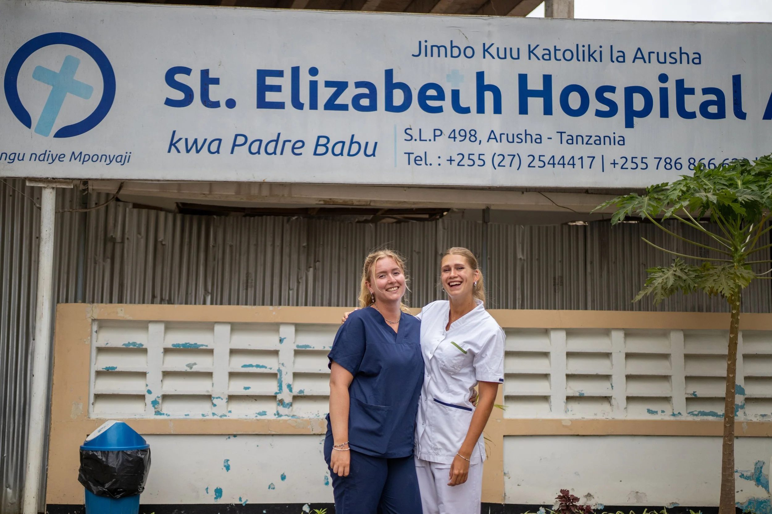 Two women, one in medical scrubs and the other in a white uniform, standing in front of a sign for St. Elizabeth Hospital in Tanzania. They are smiling with arms around each other, outdoors near a wall with a tree and a trash can nearby.