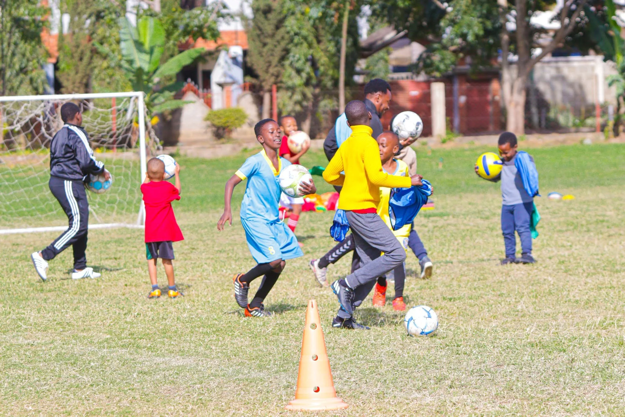 Children and a coach playing soccer outdoors on a grassy field, with a goalpost, trees, and houses in the background.