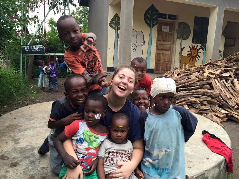 A young woman surrounded by several children smiling and posing for the camera outdoors.