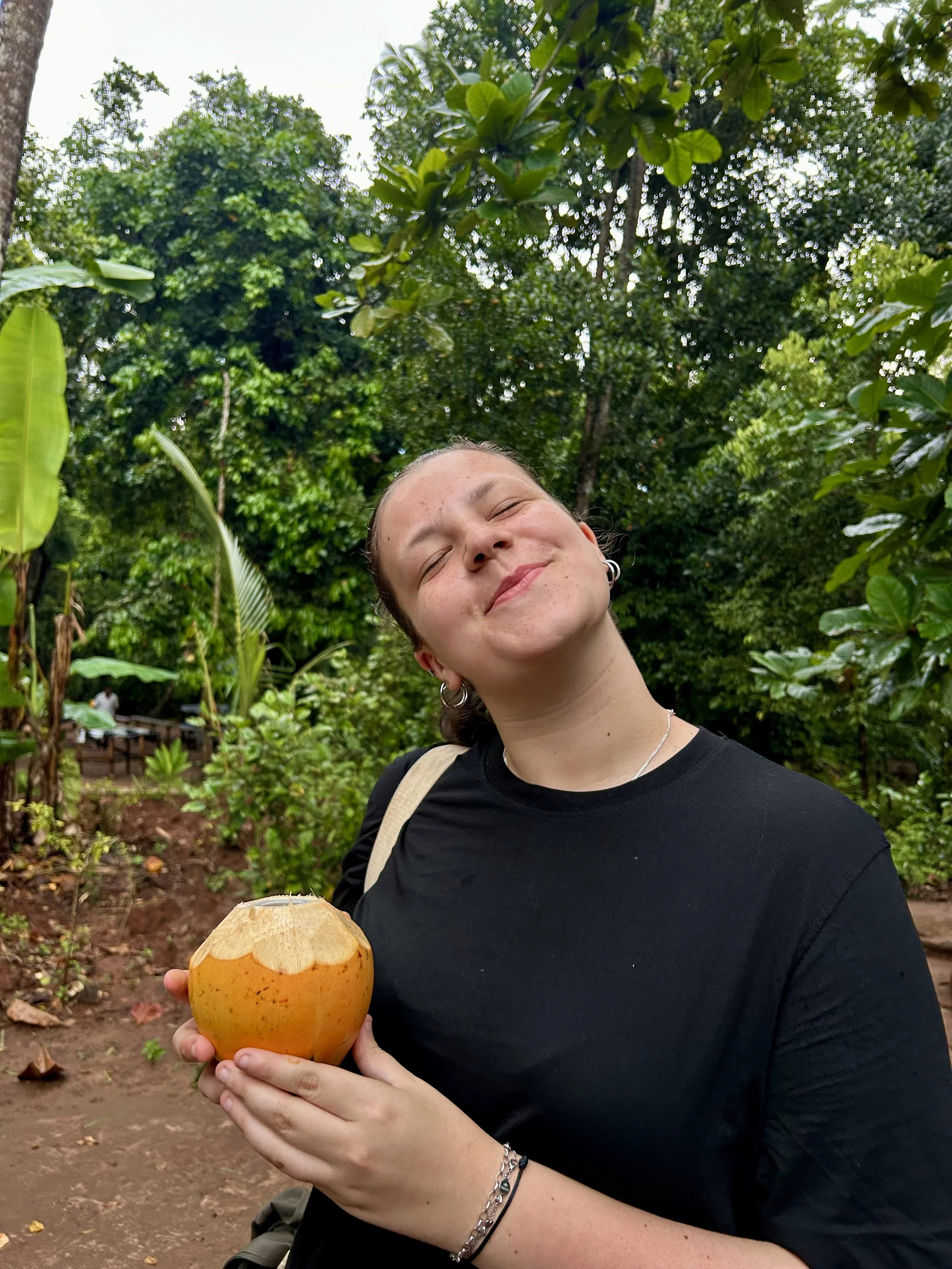 A person with light skin and short brown hair, wearing earrings and a black shirt, smiling with eyes closed, holding a large orange mushroom in a lush green outdoor setting.