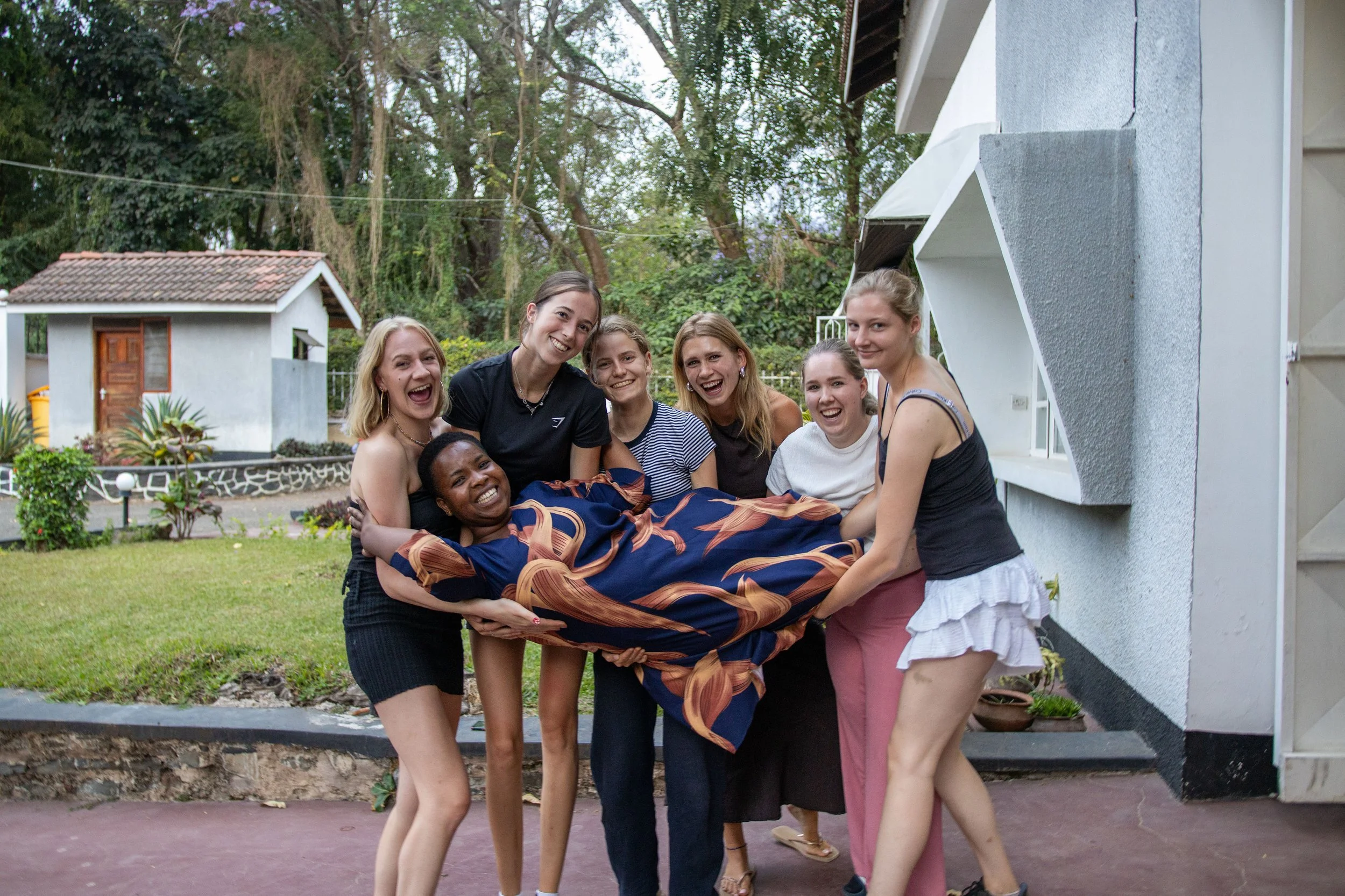 A group of seven women smiling and holding a person wrapped in a colorful blanket outdoors, with greenery and a building in the background.