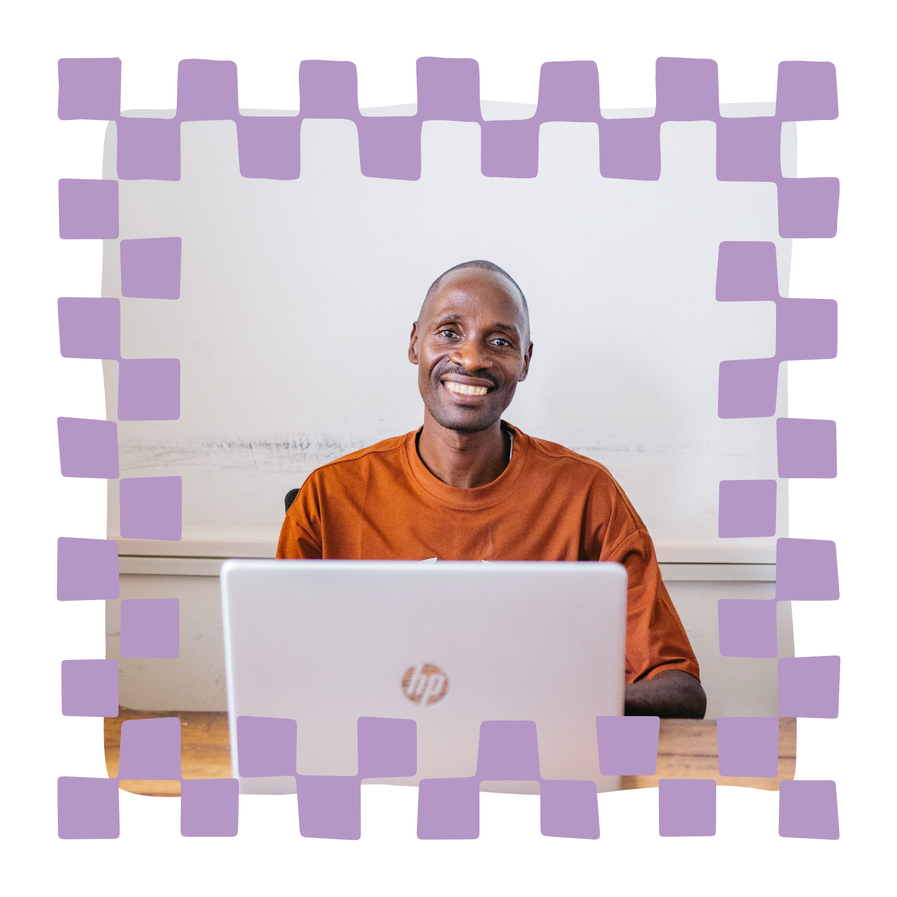 Image of a smiling man sitting at a desk with a silver HP laptop in front of him. He is wearing an orange-brown shirt and is in a room with a white wall background.