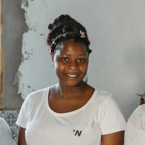 A woman with braided hair styled in an updo, smiling and wearing a white t-shirt, standing indoors against a wall.