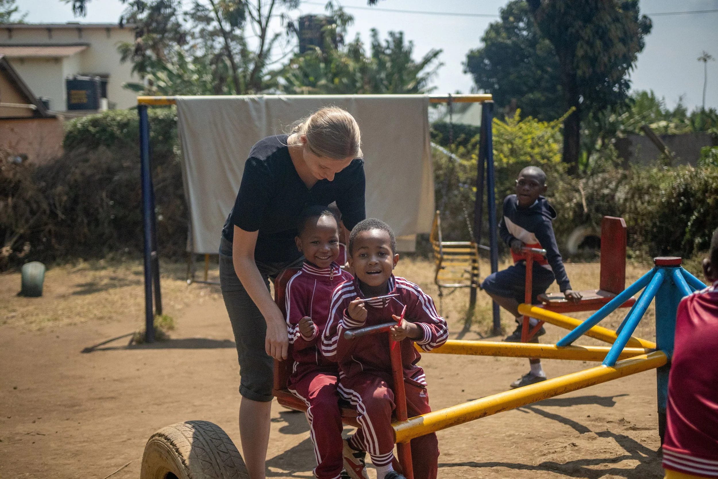A woman pushing two smiling children on a playground seesaw in an outdoor setting with trees and houses in the background.