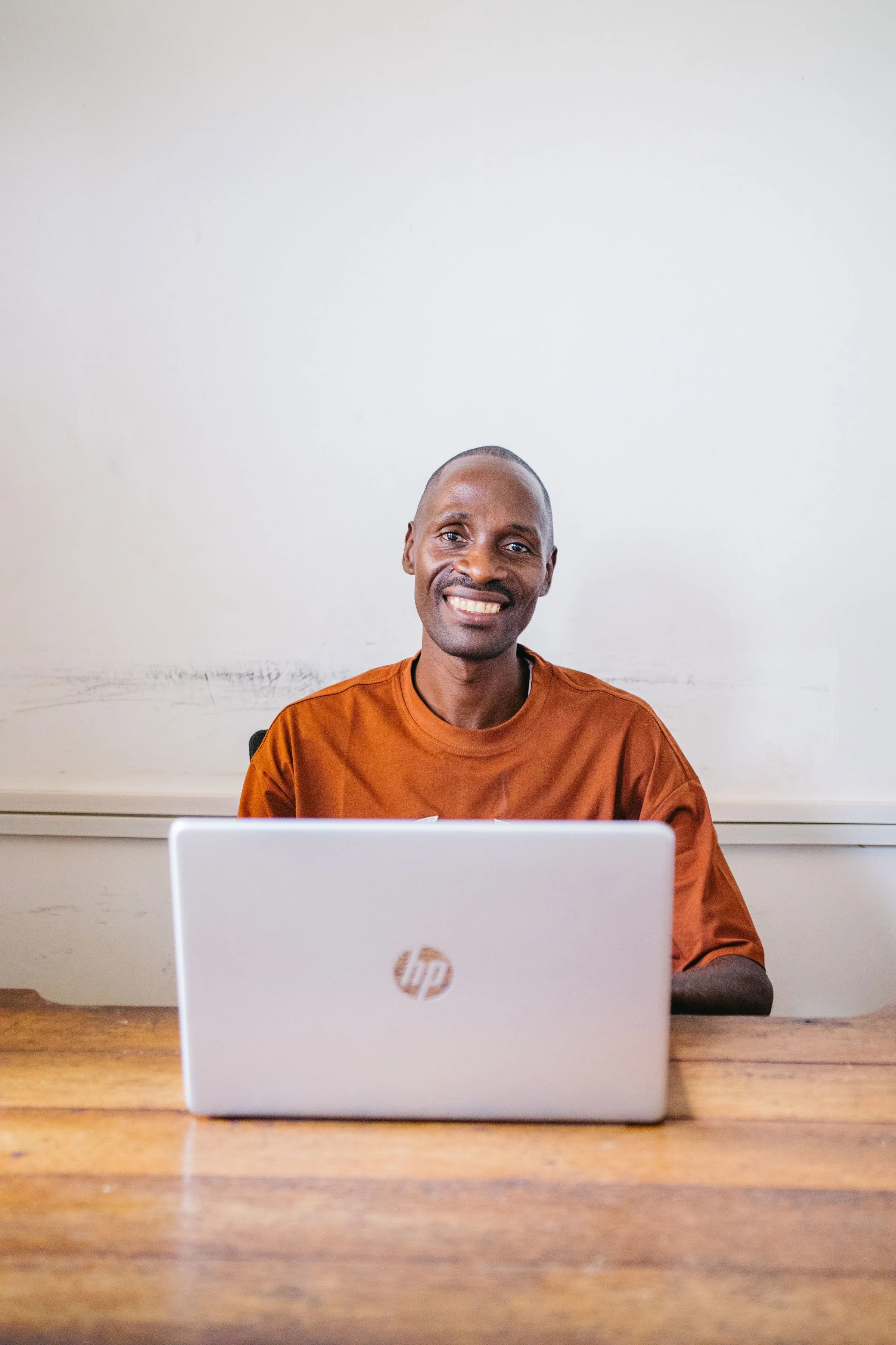 A smiling man with a shaved head wearing an orange shirt, sitting in front of a laptop at a wooden table, with a plain white wall in the background.