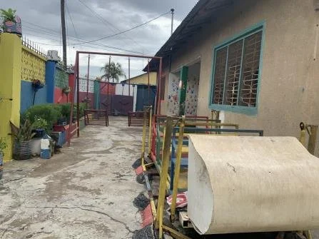 A courtyard area with a concrete path, colorful fences, a small building with large windows, plants, a trash bin, and a large rolled-up object.