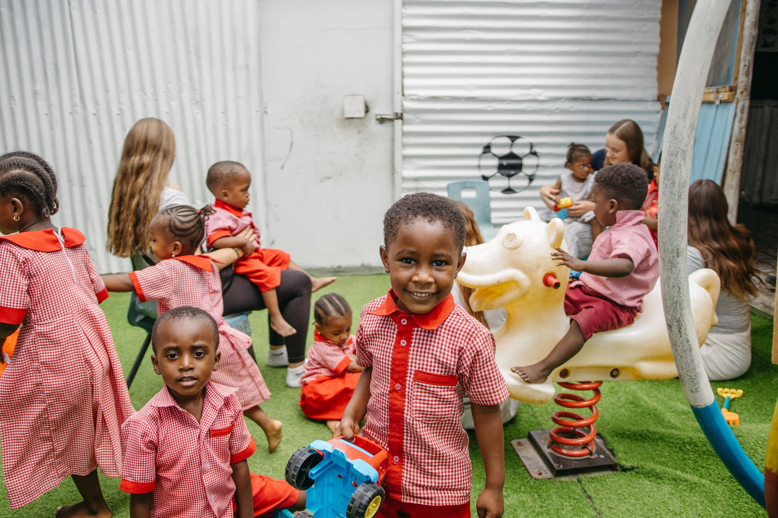 Children playing outside in a playground area with grass and metal and plastic equipment, wearing red and white checkered uniforms, with a woman supervising.
