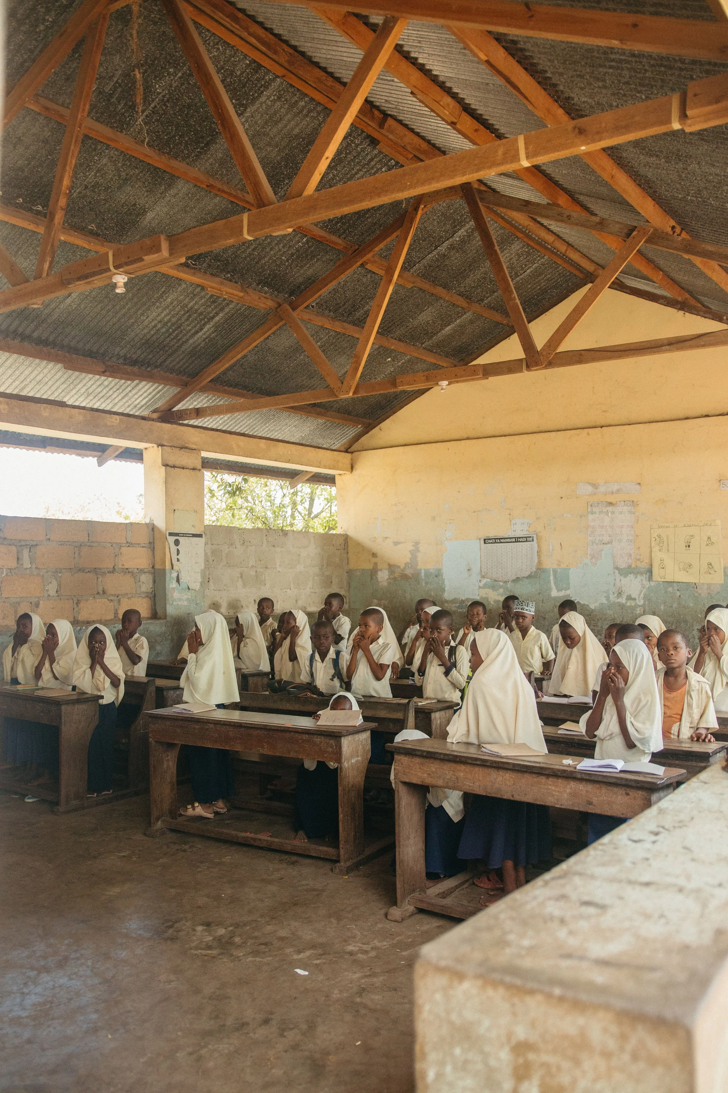 A classroom with students wearing white uniforms, sitting at wooden desks, and raising their hands. The classroom has a concrete floor, unfinished brick walls, and a ceiling with wooden beams and corrugated metal roofing.