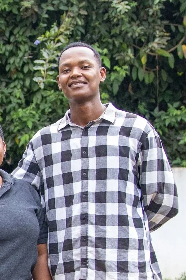 Young man smiling outdoors with green foliage background, wearing a black and white checkered shirt.
