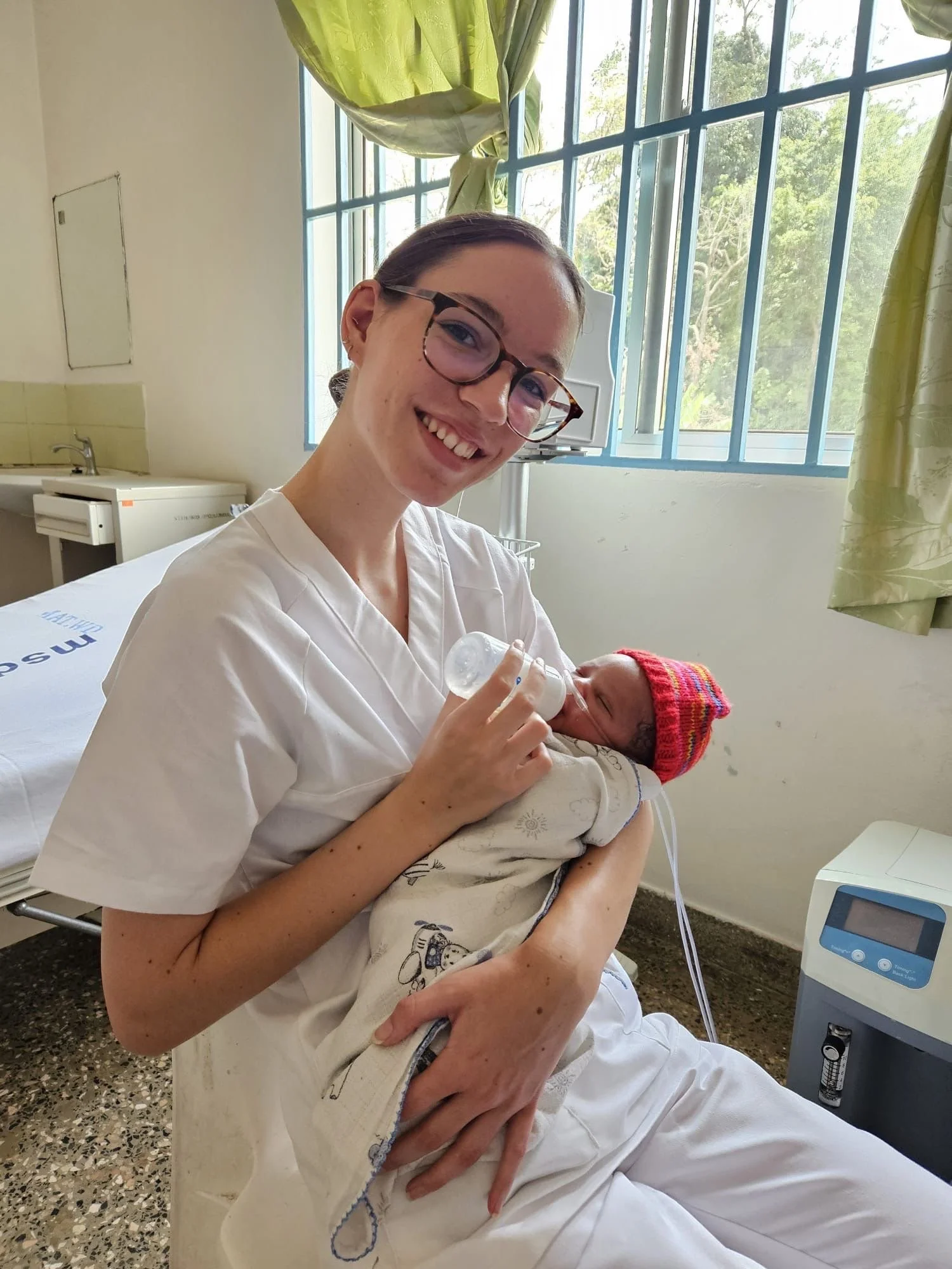 A woman in white scrubs is smiling while holding a newborn baby wrapped in a blanket with her left arm; the woman appears happy and the baby is wearing a colorful knit hat and is being fed with a bottle.