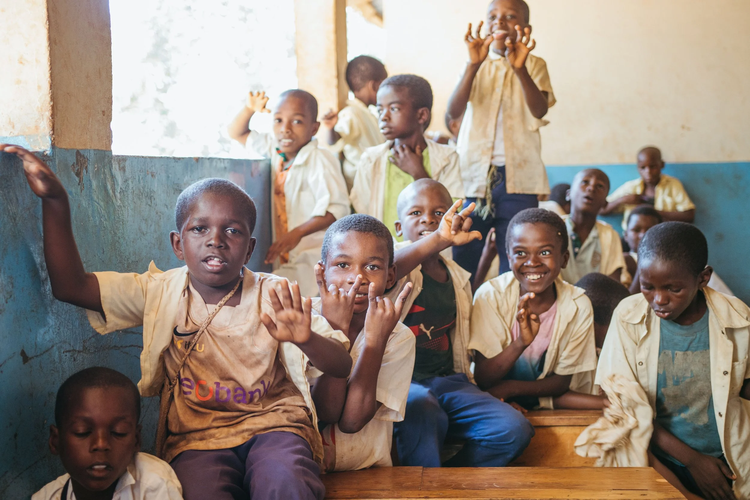 A group of children sitting and standing on indoor wooden steps, smiling and making gestures. They wear light-colored shirts, some with text or logos, in a classroom with a blue and yellow wall and a window letting in natural light.