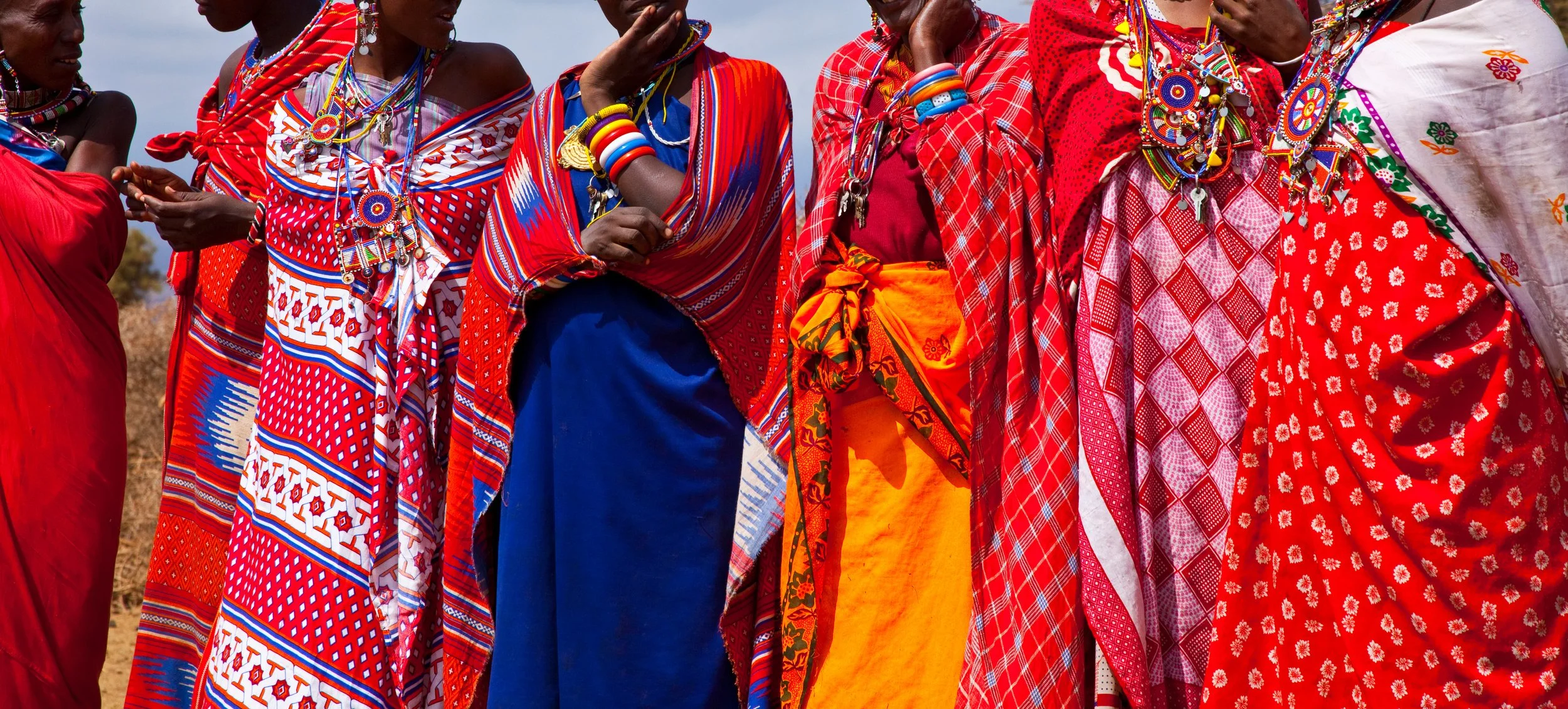 Group of African women dressed in colorful traditional clothing and jewelry, standing outdoors.