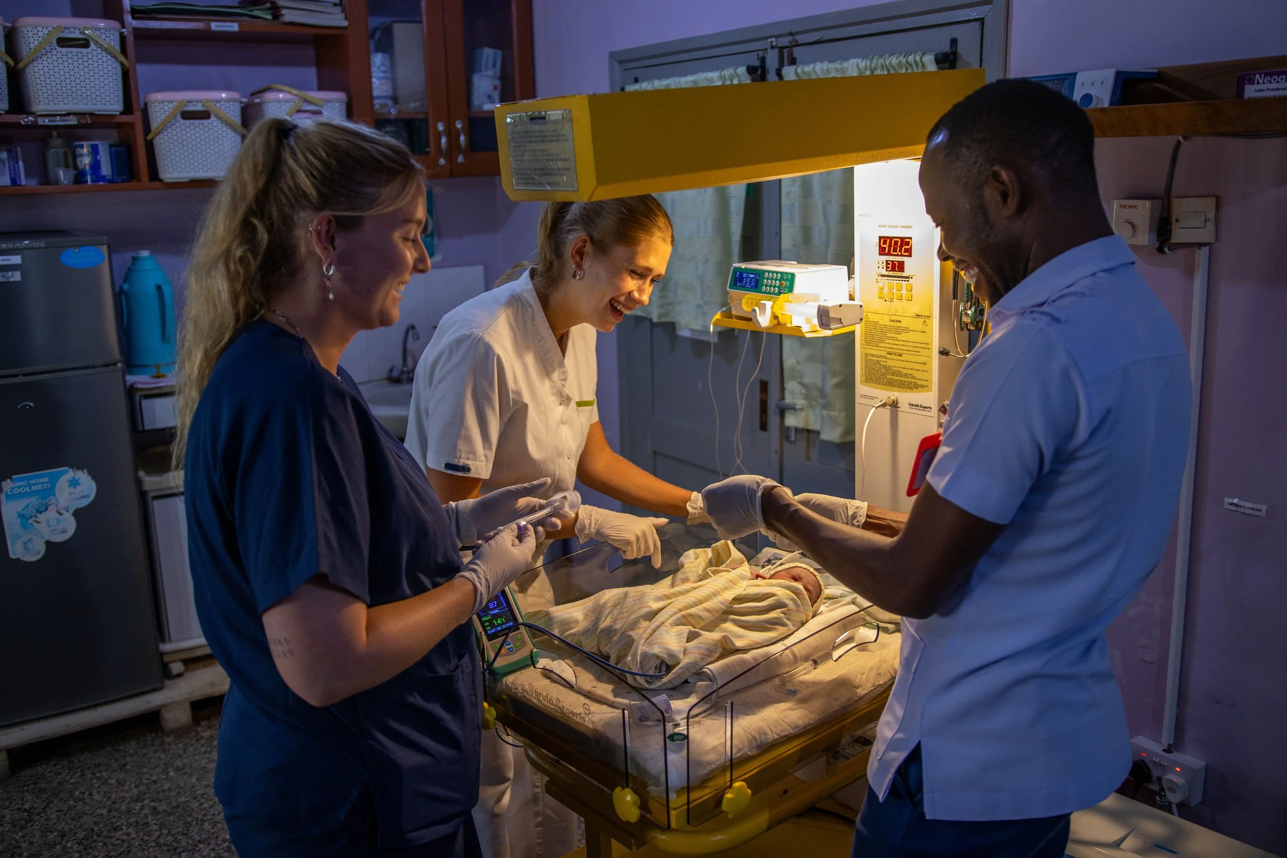 Medical staff attending to a newborn in a hospital room with three staff members, medical equipment, and a hospital bed.