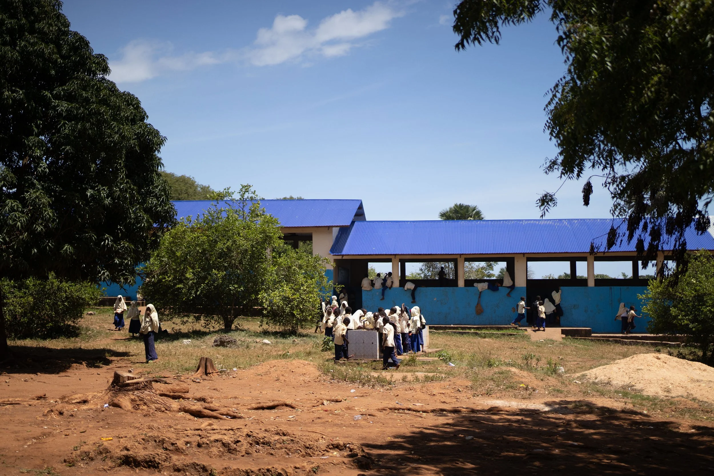 A schoolyard with students in uniforms gathered outside a building with a blue roof, surrounded by trees under a partly cloudy sky.