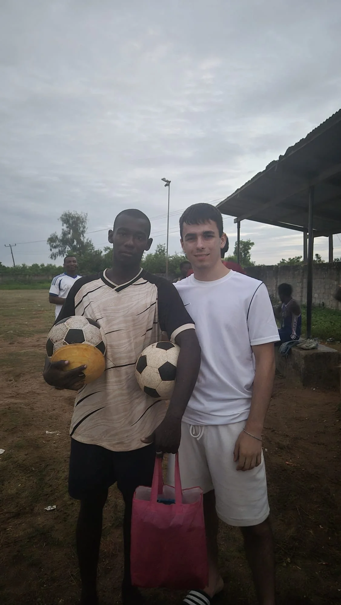Two young men standing outdoors on a cloudy day, holding soccer balls and a yellow ball, with other people in the background, one wearing a white shirt and shorts and the other in a patterned shirt.