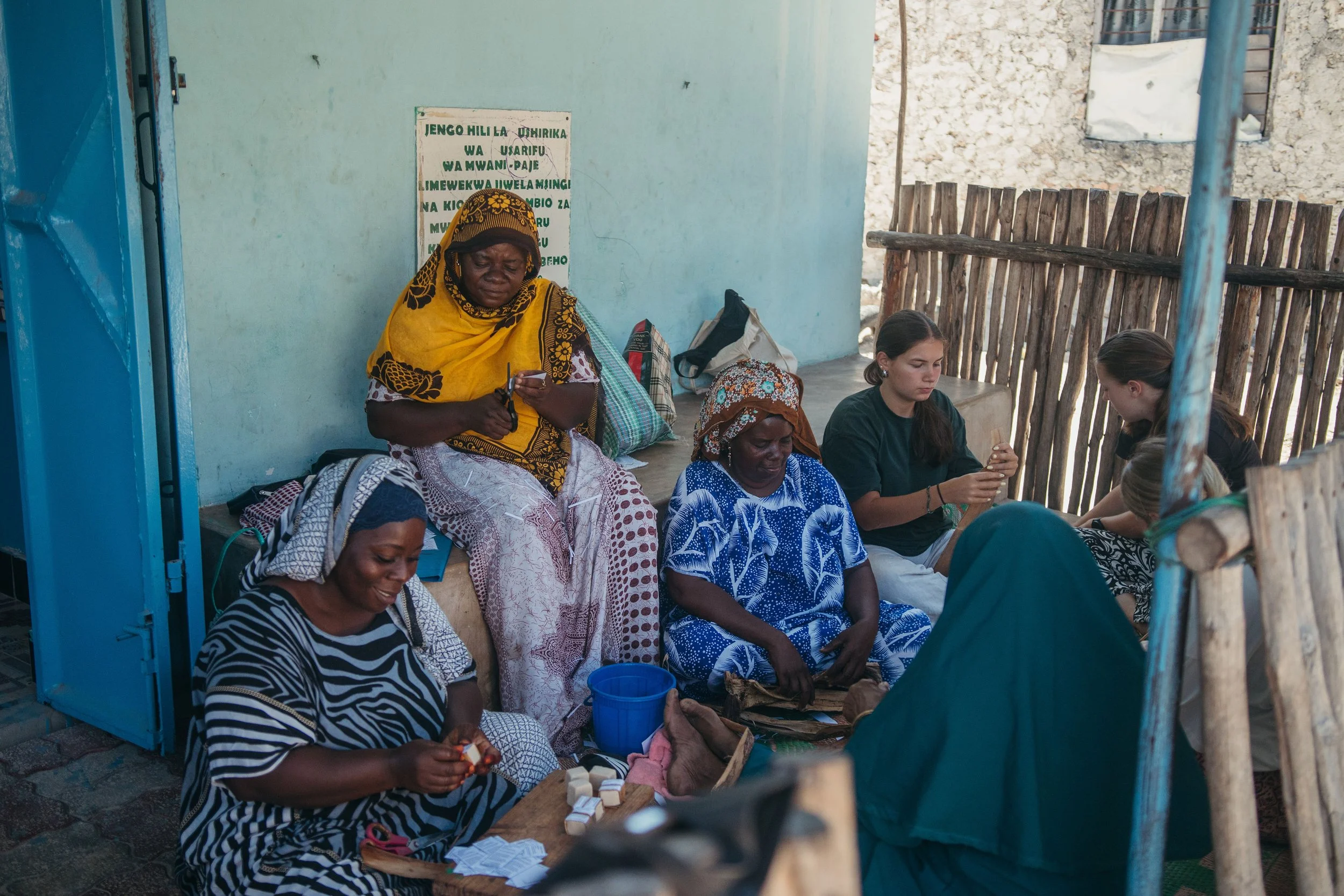 Group of women sitting and working together outdoors, with a light blue wall and wooden fence in the background.
