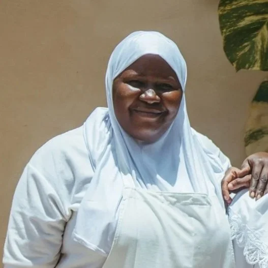 A smiling woman wearing a white hijab and dress, standing outdoors with a beige wall and green plant in the background.