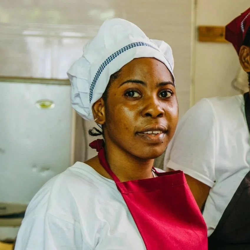 A woman wearing a white chef's hat and apron with a red necktie, standing in a kitchen.