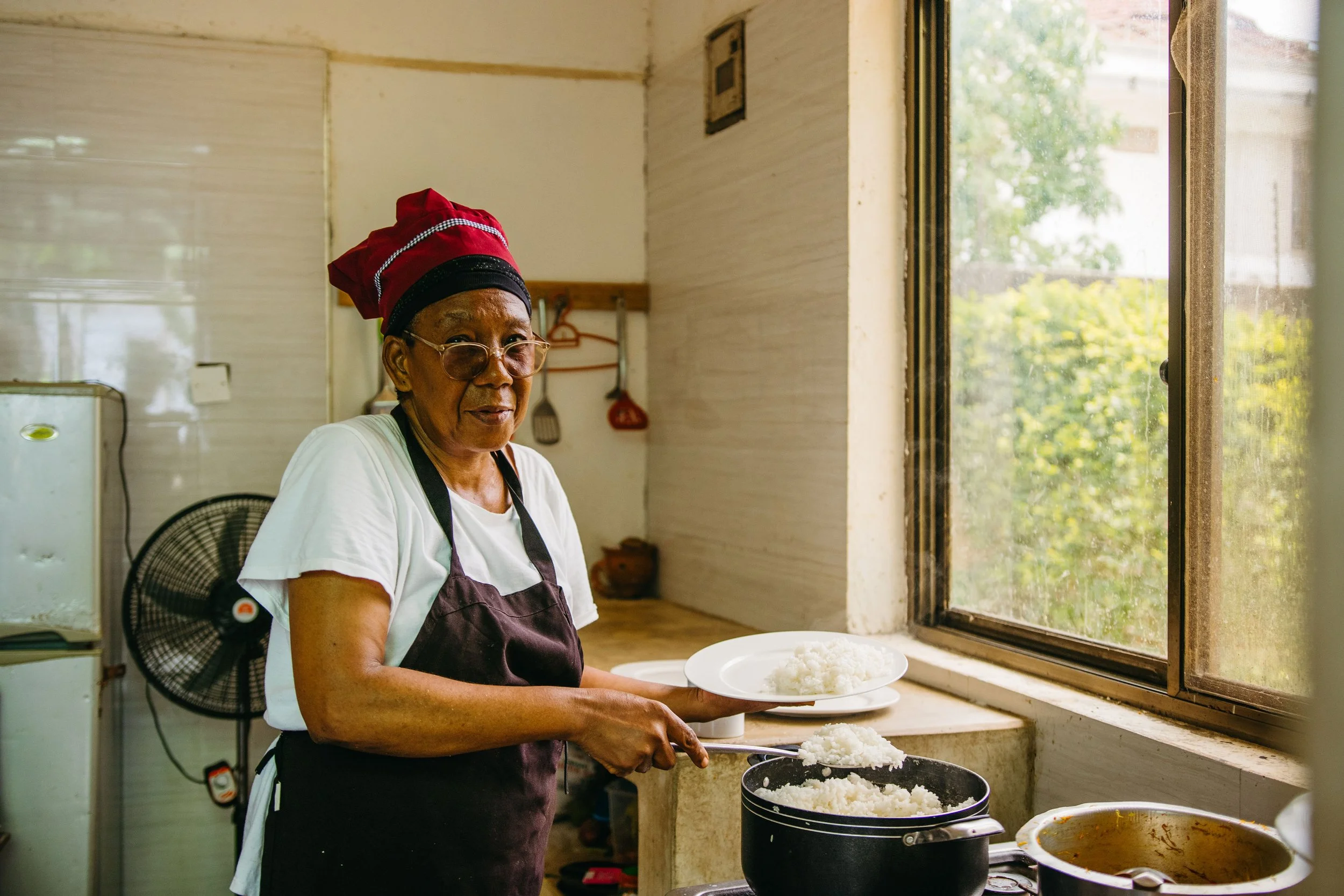An elderly woman wearing glasses and a red chef hat is serving rice from a pot in a kitchen next to a window with greenery outside.