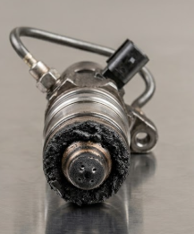 A car fuel injector with a black nozzle and metal body placed on a metallic surface