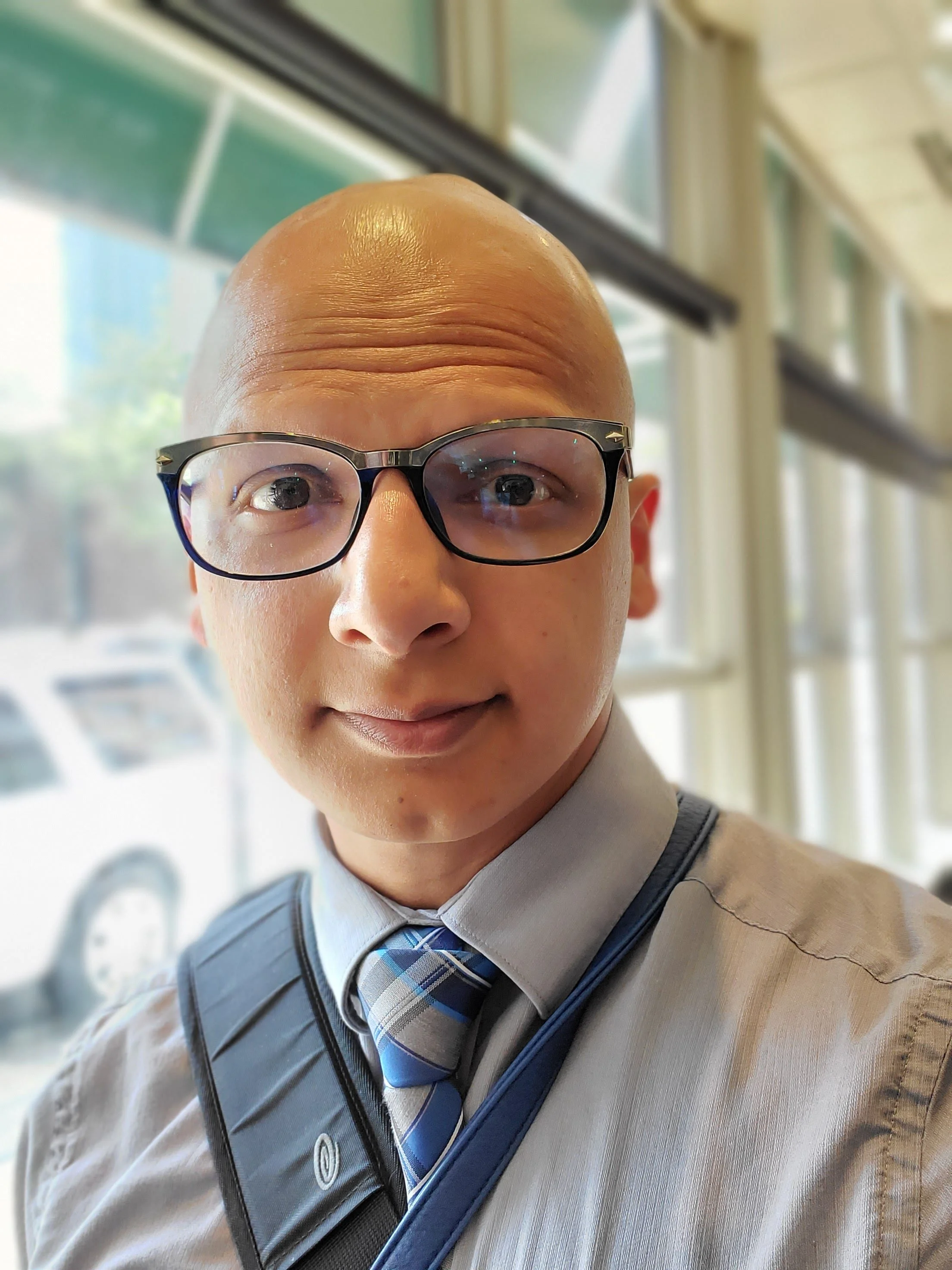 Close-up of a person with a shaved head, wearing glasses, a light-colored shirt, and a blue plaid tie, in a bright indoor setting with windows.
