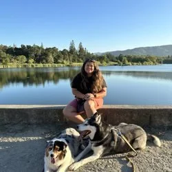 A woman sitting on a stone ledge by a lake with three dogs, surrounded by trees and mountains under a clear blue sky.
