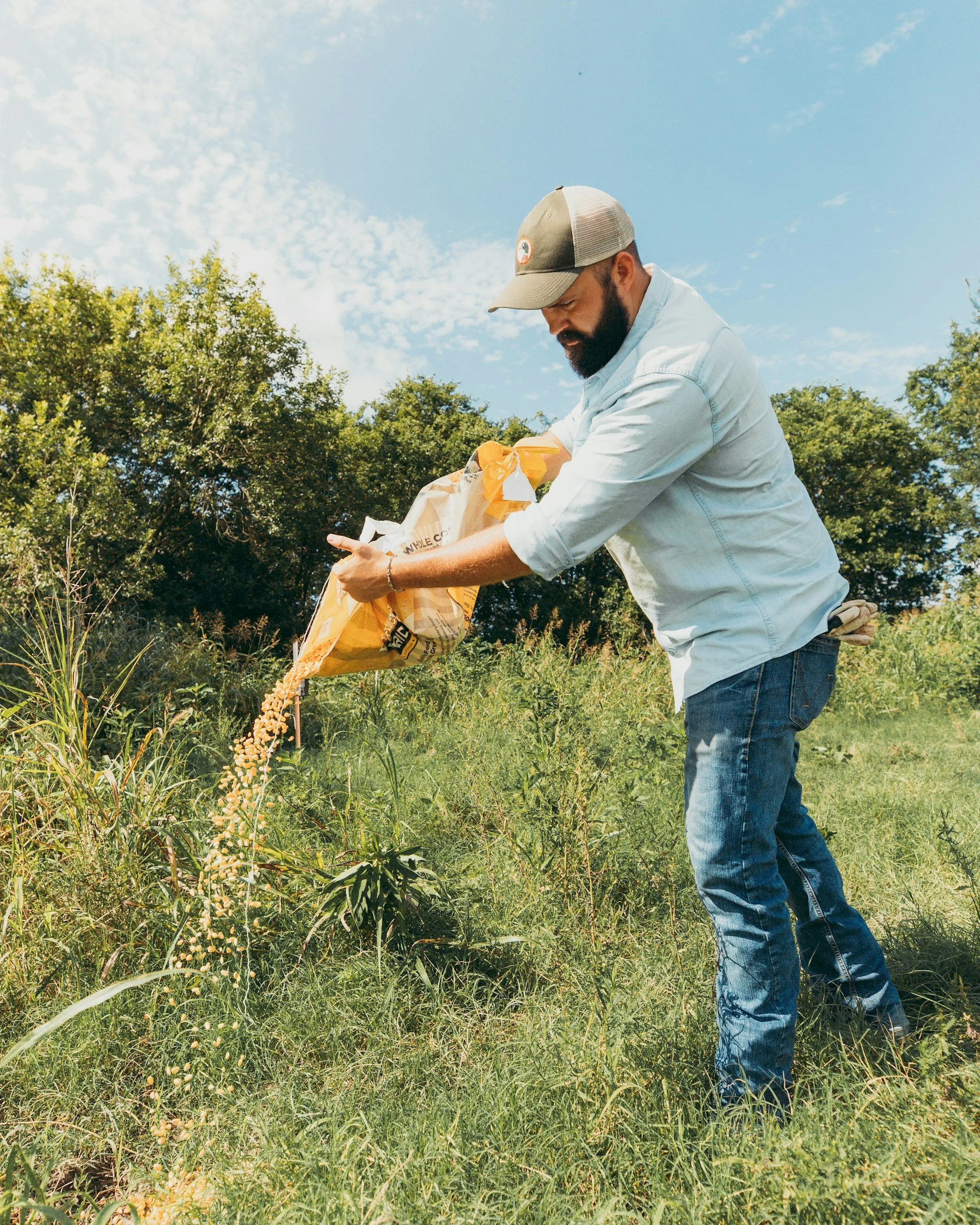 Man in casual clothes spreading seeds from a bag in a field with green grass, trees, and a partly cloudy sky.