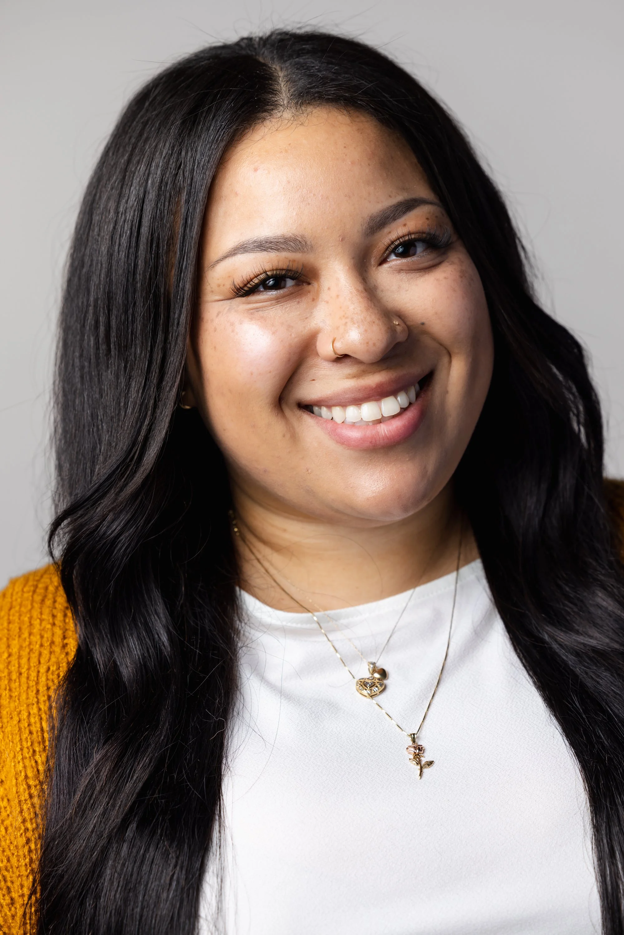 Photo of Pyrite & Pearl owner, Chai. She has long, black hair, is smiling at the camera, wearing a white shirt with an unbuttoned mustard yellow cardigan, and layered gold necklaces with heart and rose pendants.
