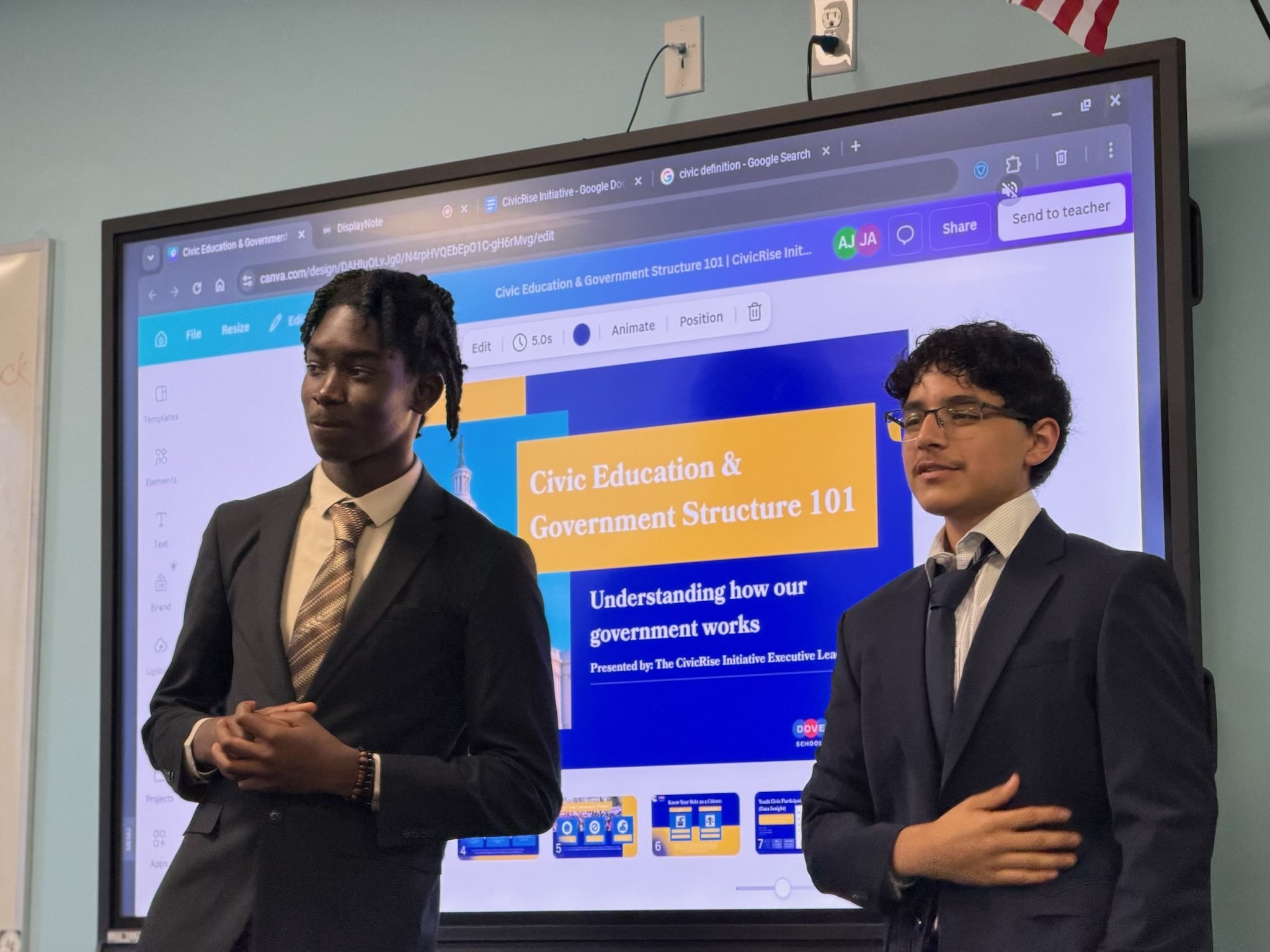 Two young men in suits standing in front of a large screen displaying a presentation titled "Civic Education & Government Structure 101." One man has dreadlocks and the other has curly hair with glasses. The screen shows a webpage with menu options and colorful graphics.