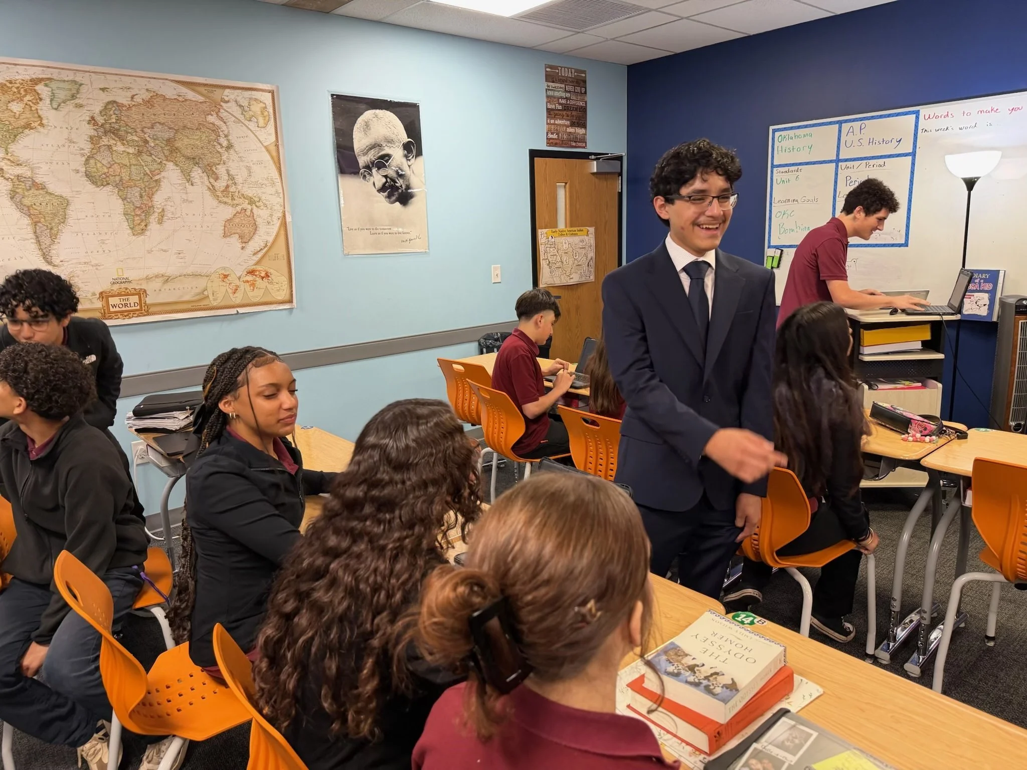 Classroom with students, some wearing uniforms, seated at tables, and two students standing at the front, one smiling and one working on a laptop. The room has educational posters, a world map, and a whiteboard.