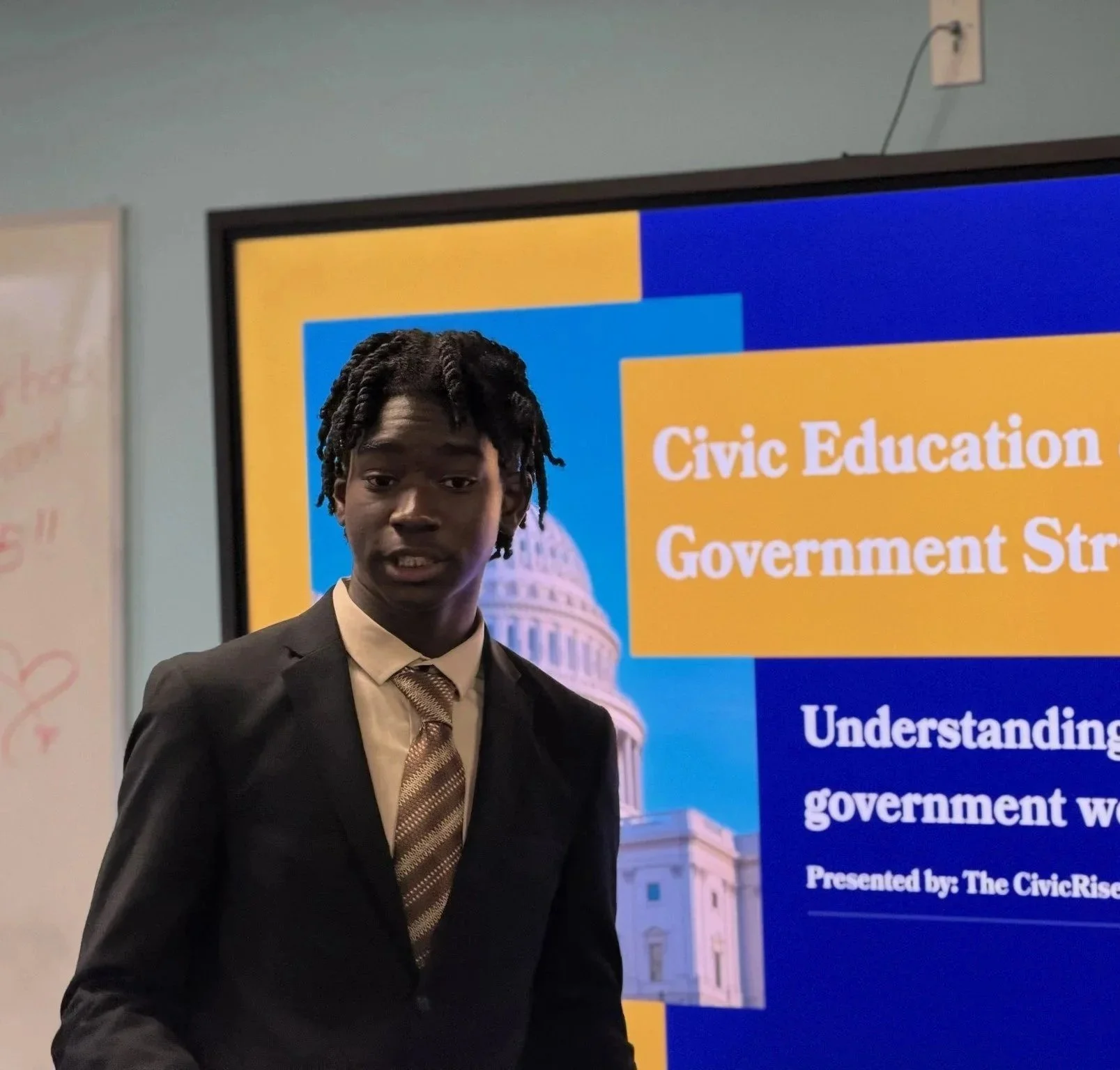 CivicRise Initiative Founder and Executive Director Jakub Animashaun in a suit standing in front of a presentation slide about civic education and government strategy, with part of the U.S. Capitol building visible in the background.