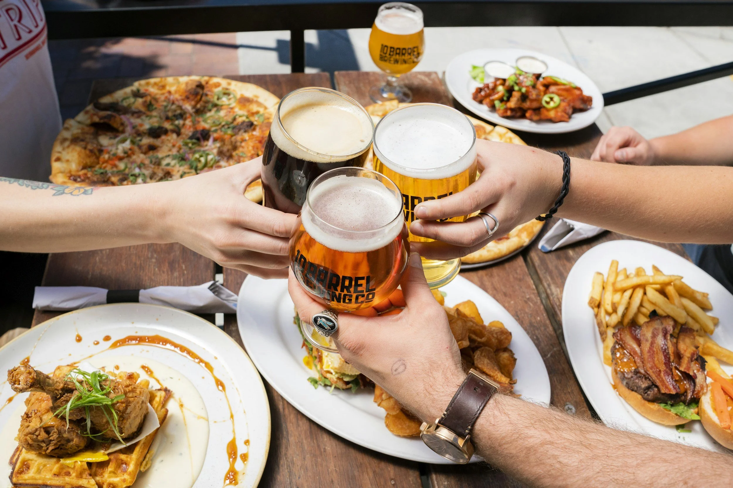 People clinking glasses of beer over a table filled with pizza, fried chicken, and other dishes at an outdoor restaurant.