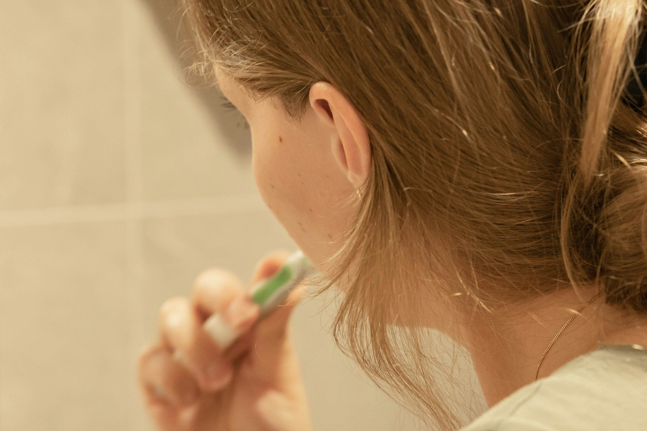 A woman with light brown hair and a gold necklace is seen from behind, holding a small mirror or compact close to her face.