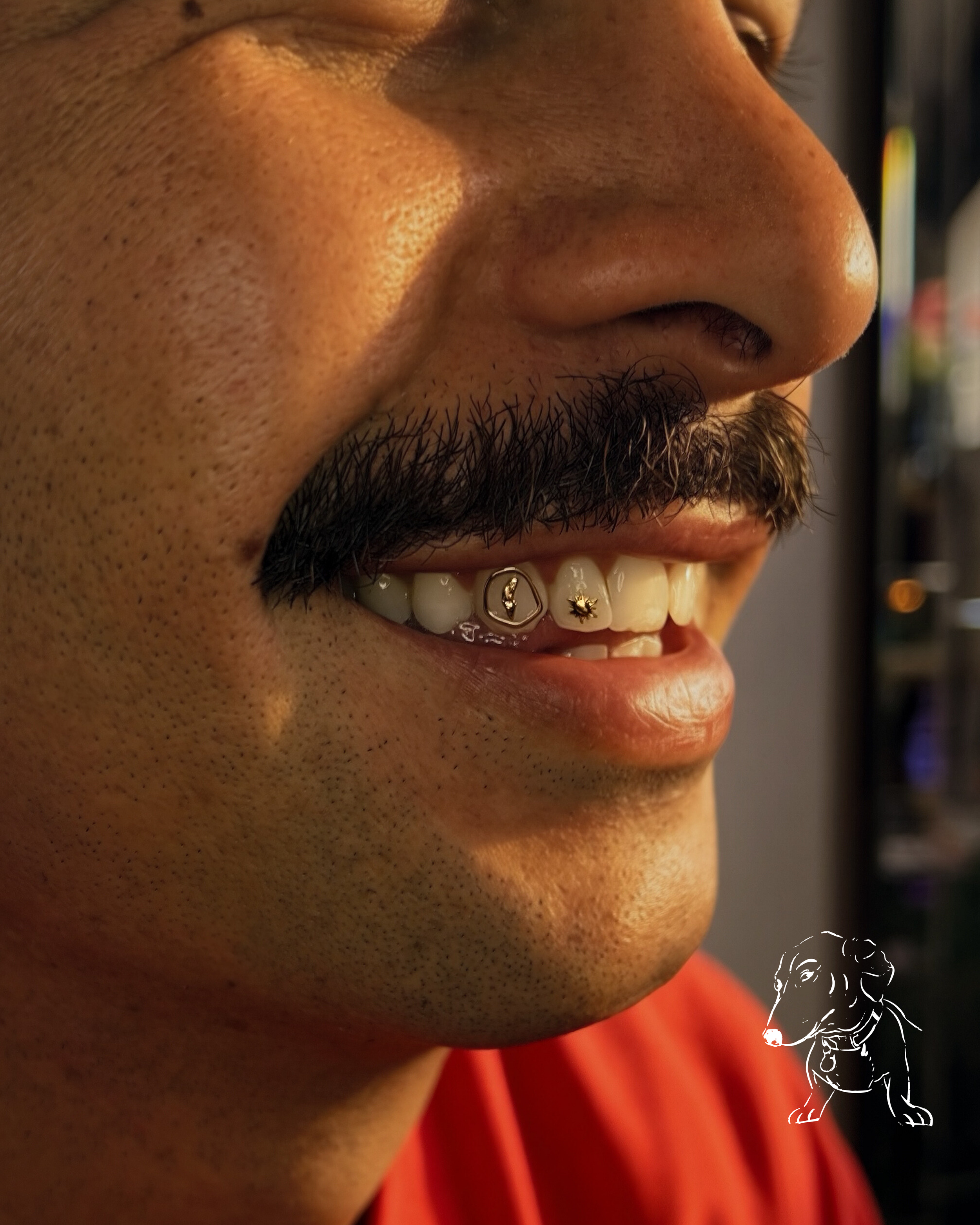 Close-up of a person's face showing their nose, mustache, and lips, with a smile revealing teeth. The person has dark hair and is wearing a red shirt. A small, white line-drawing of a dog with a ball in its mouth is present in the bottom right corner.