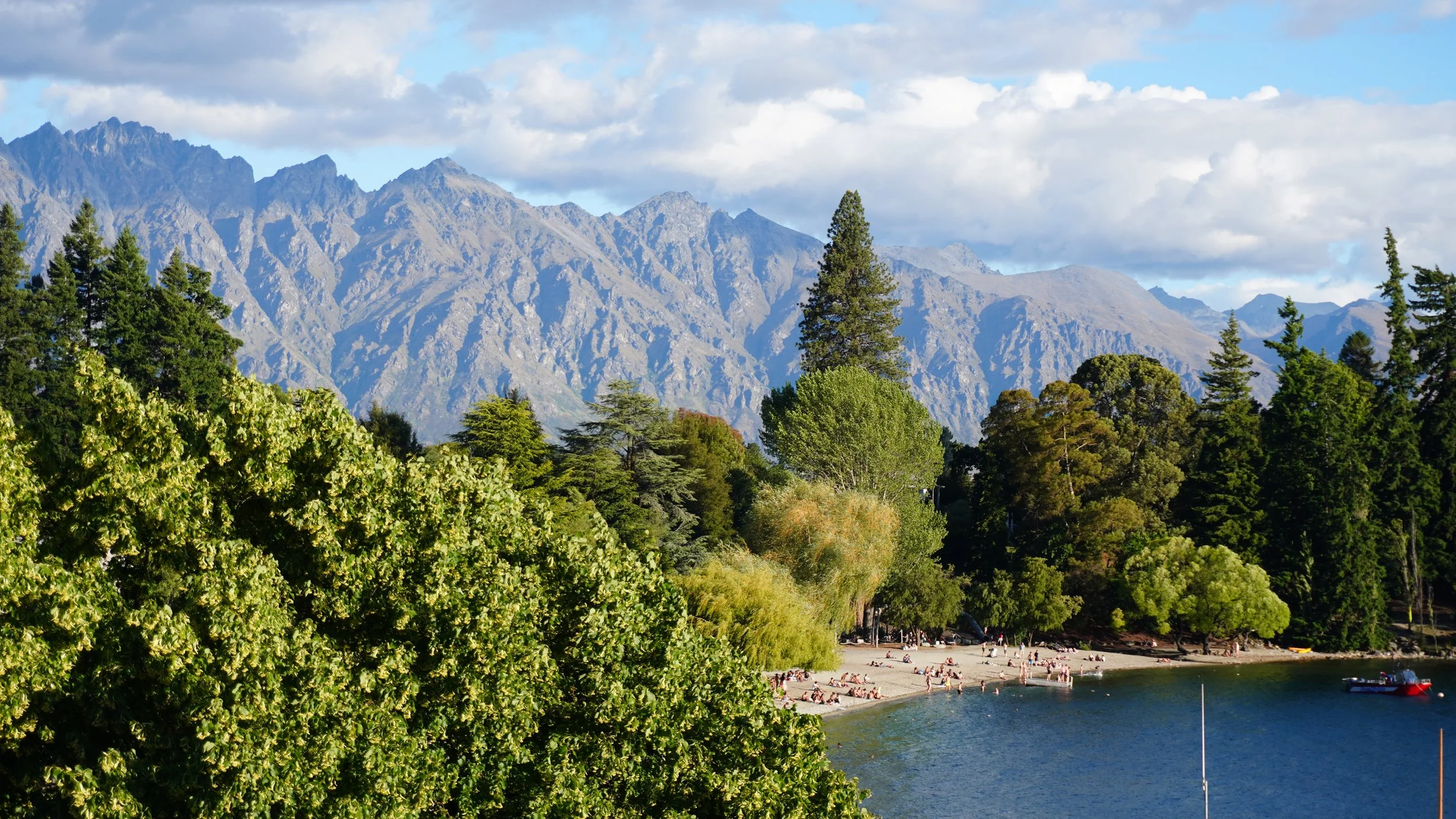 Scenic view of a lake with a crowded beach, surrounded by lush green trees and tall mountains in the background under a partly cloudy sky.