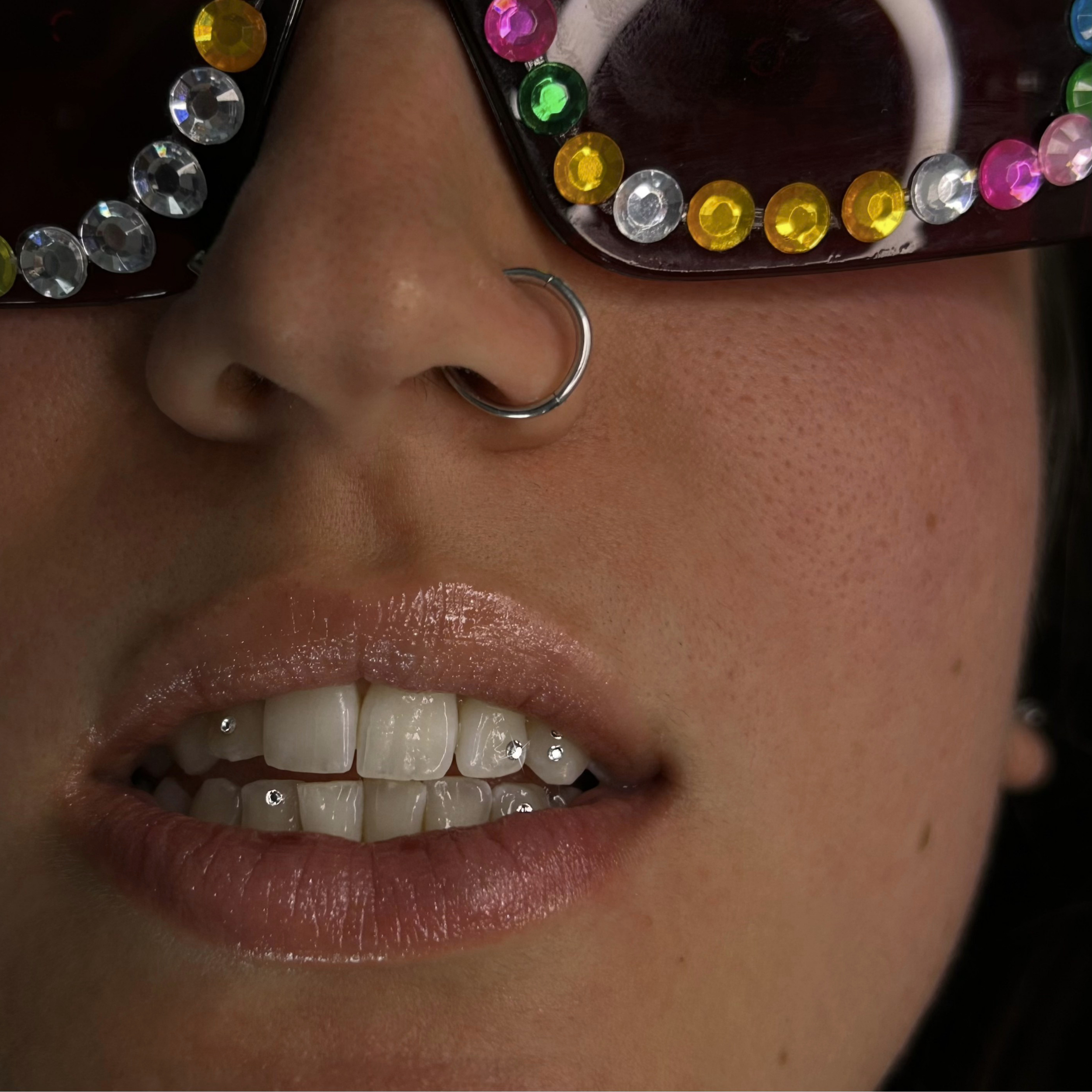 Close-up photo of an individual wearing sunglasses decorated with colorful rhinestones, showing part of the nose and mouth with piercings.