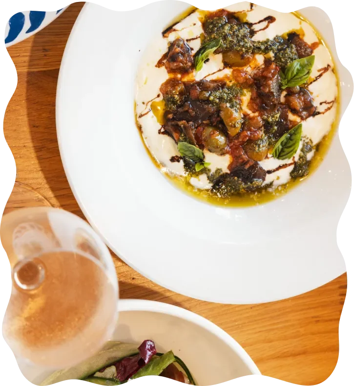 A plate of burrata cheese topped with basil, olive oil, and balsamic glaze, served with a glass of beer and a side salad.