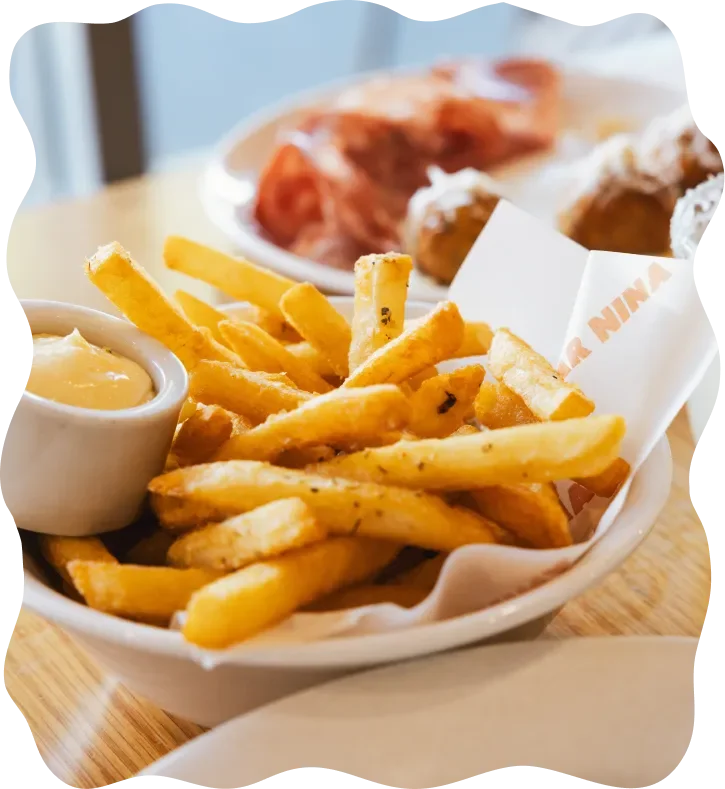 French fries in a basket with a side of dipping sauce, likely cheese or gravy, and a plate of fried chicken in the background.