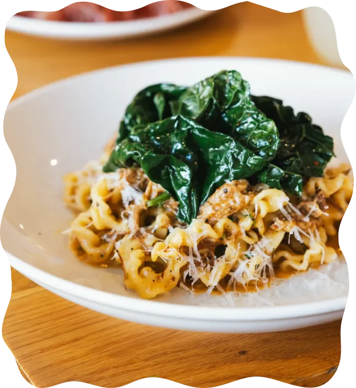 A bowl of cooked pasta topped with shredded cheese and dark green leafy vegetables, possibly spinach.