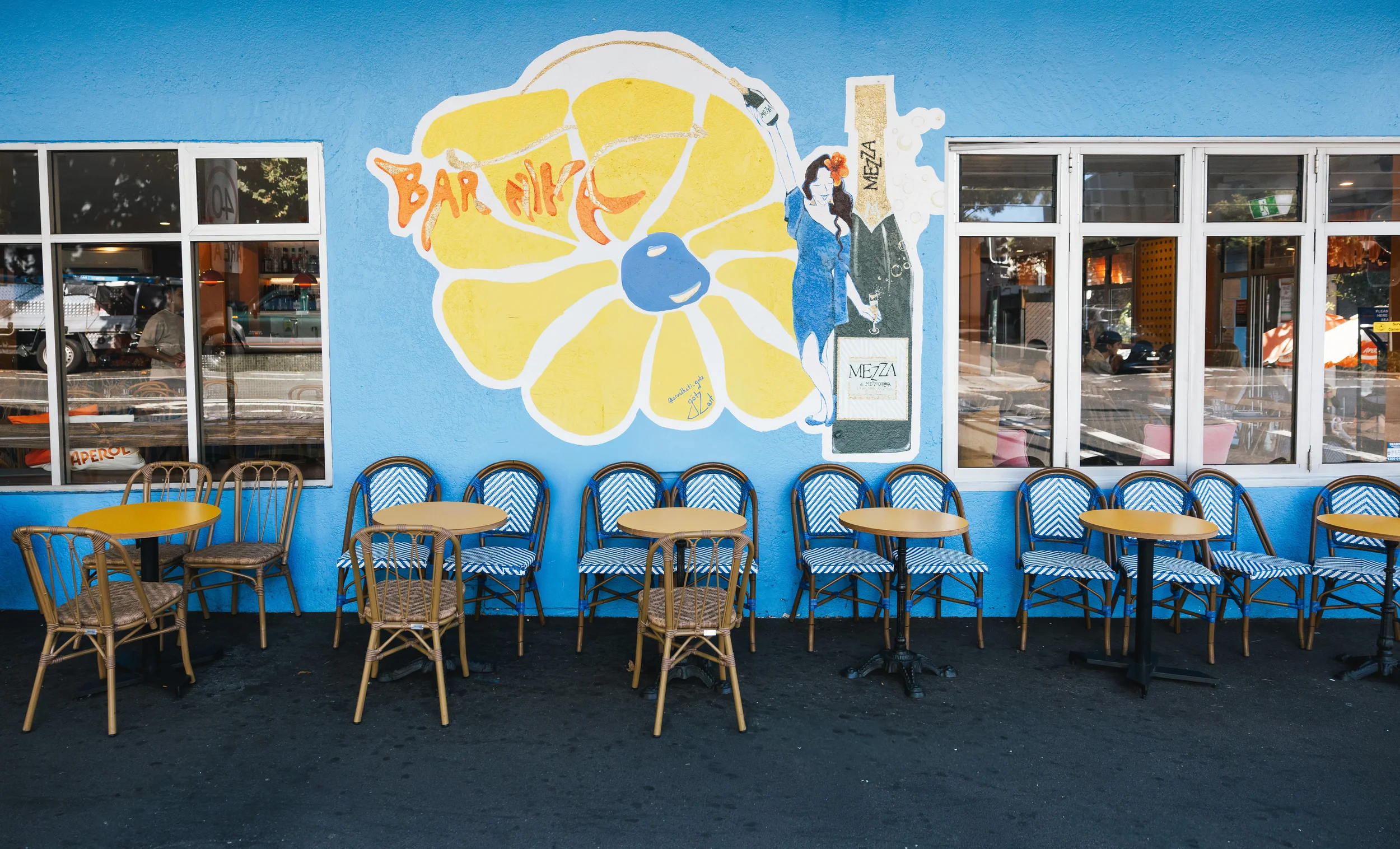 Colorful mural of a woman holding a beer and a large lemon slice, with the words 'Bar Nica' and a bottle of Mezza wine, outside a café with empty blue chairs and yellow tables.