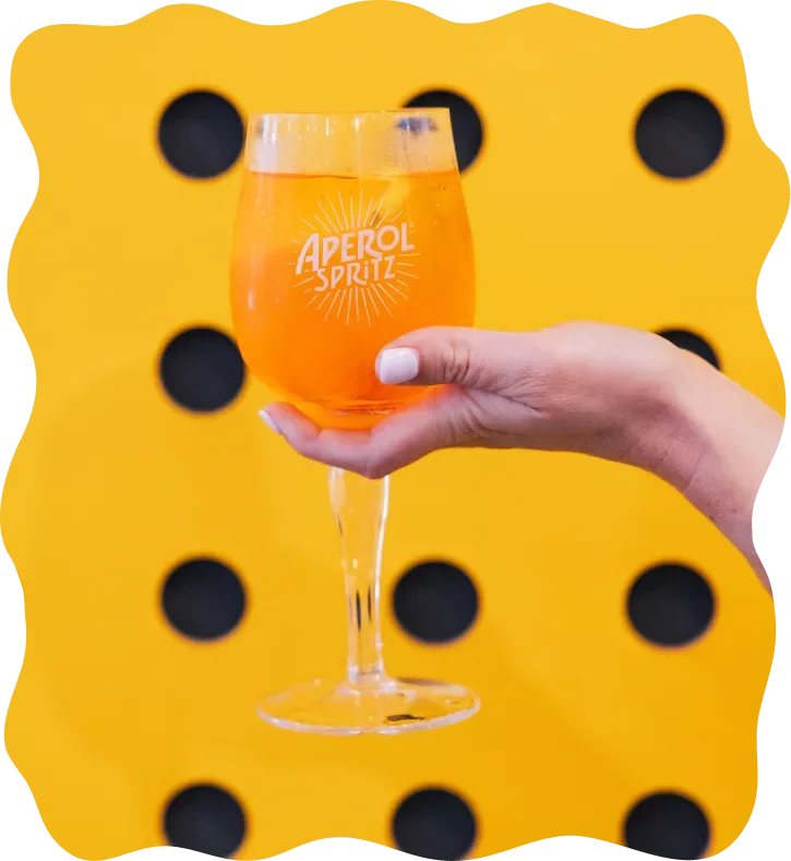 A hand holding a glass of Aperol Spritz in front of a yellow background with black circular cutouts, resembling a slice of cheese.