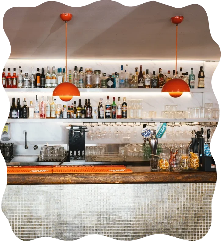 Bar counter with bottles of alcohol, glasses, and bar tools, illuminated by two orange pendant lights.