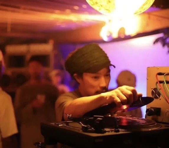 A young man is operating turntables as a DJ. The background shows a nighttime scene of a club or event venue with people and colorful lights