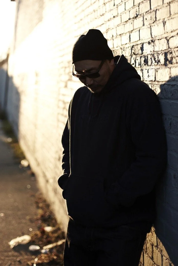 A man wearing a black hat and sunglasses is leaning against a white brick wall. The setting sun casts shadows over his face and body