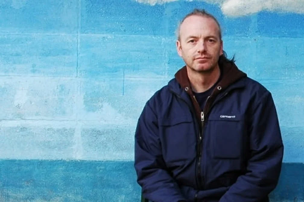 A photograph of a man sitting in front of a blue and white wall