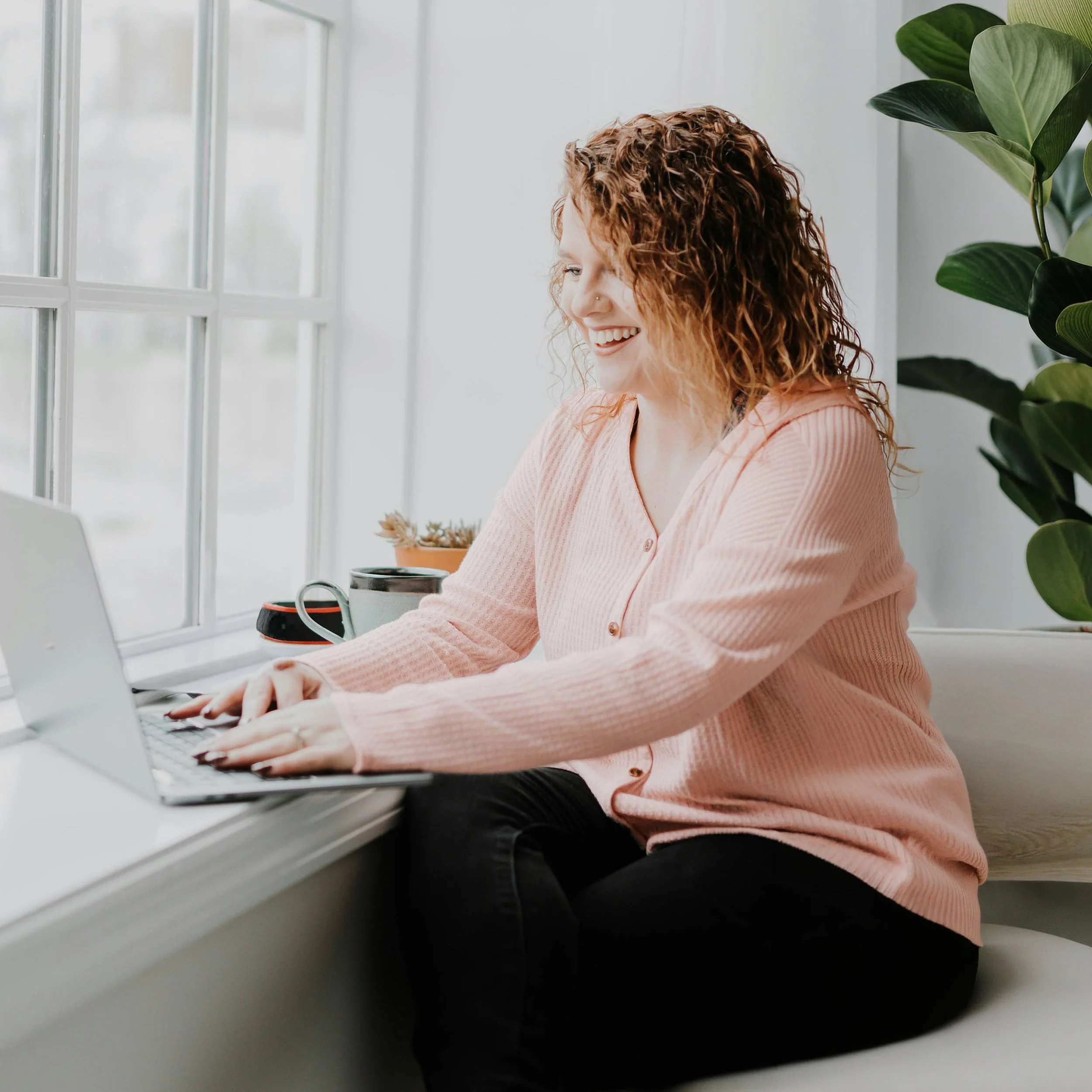 A woman with curly red hair and a nose piercing working on a laptop at a desk beside a window and a large green plant.